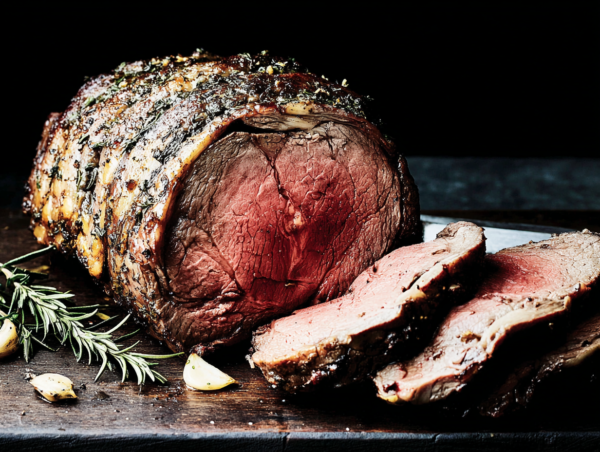 Beef Fillet Whole with a pink centre is sliced on a wooden board, served beside herbs and garlic cloves.
