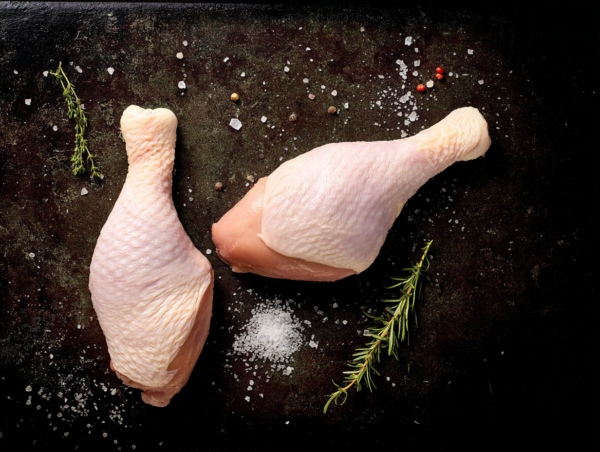 Two Free Range Chicken Legs on a dark surface with rosemary, peppercorns, and coarse salt scattered around.