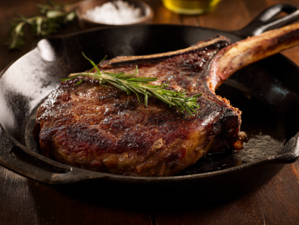 A cooked Cowboy Steak with rosemary in a cast iron frying pan on a wooden table.