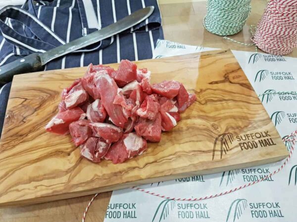 Diced lamb on a wooden board with a knife, string, and Suffolk Food Hall branded paper nearby.