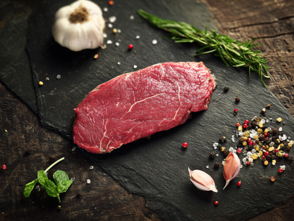 Minute steak on a slate board with garlic, rosemary, mint, salt, and peppercorns on a wooden table.
