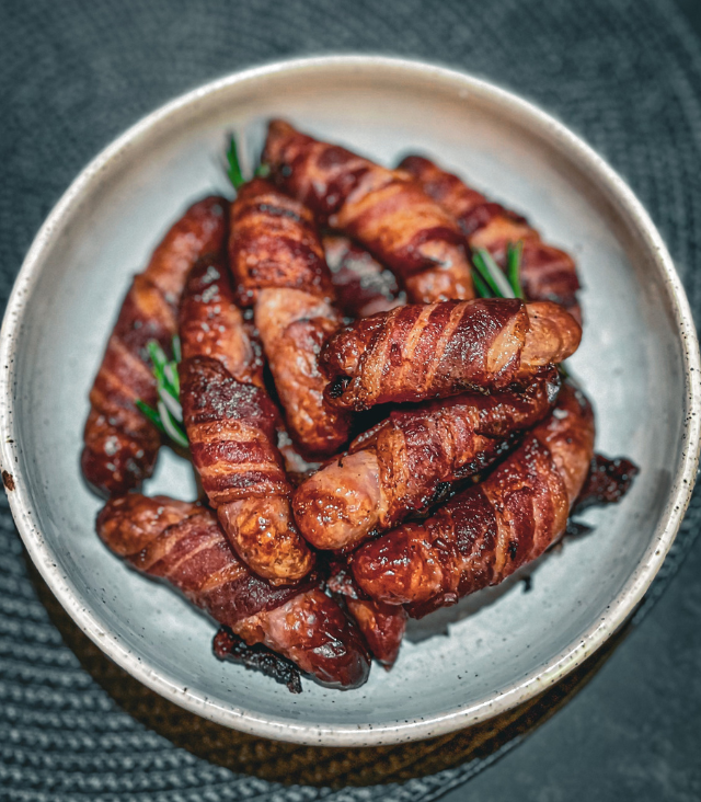 A bowl of pigs in blankets, cooked and garnished with sprigs of rosemary.