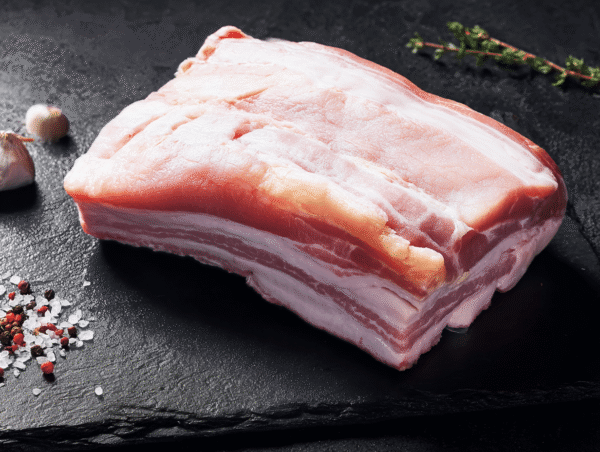 Pork belly on black slate with herbs, garlic, salt, and peppercorns nearby.