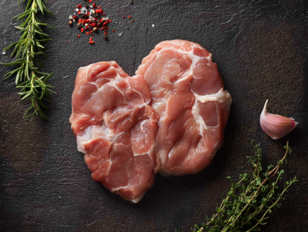 Two Pork Shoulder Steaks on a dark surface with herbs, peppercorns, and a garlic clove nearby.