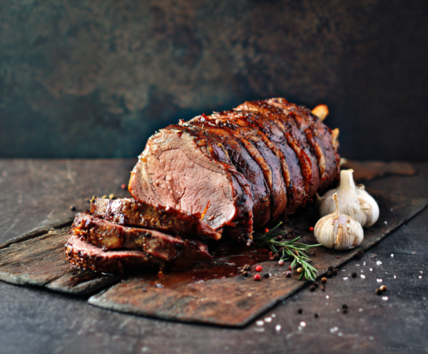 Rolled Beef Sirloin sliced on a wooden board with garlic, rosemary, salt, and peppercorns.