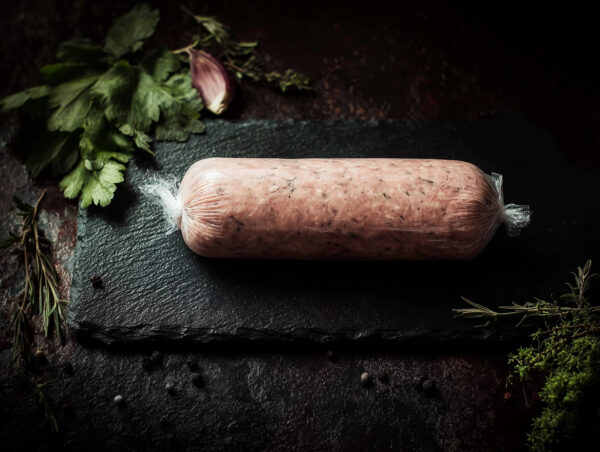 Sausage meat log on a slate board, surrounded by fresh herbs and peppercorns.