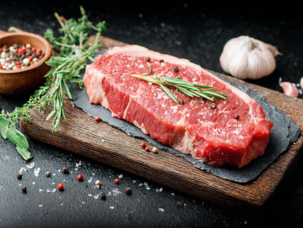 Sirloin steak on a wooden board with rosemary, sea salt, peppercorns, and garlic in the background.