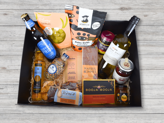 The Ultimate Hamper (NEW) with wine, beer, snacks, chocolates, preserves & coffee on a wooden surface.