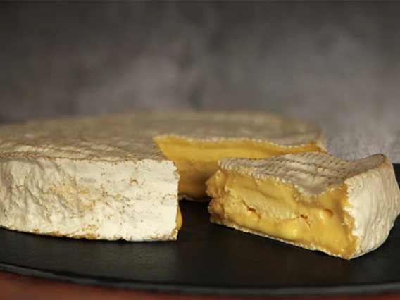 A Suffolk Brie wheel with a wedge cut out reveals its creamy interior on a dark surface.