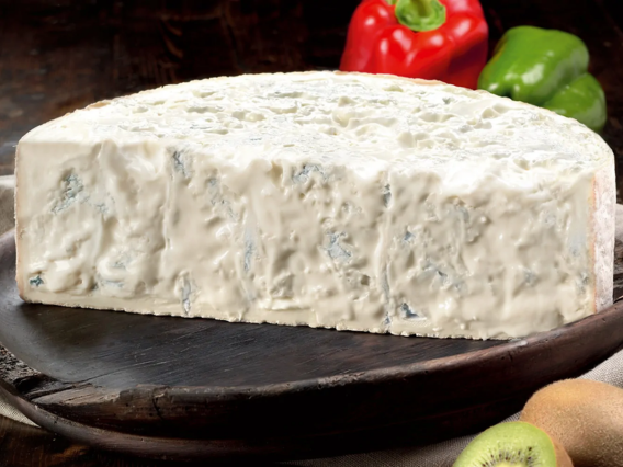 A large wedge of Gorgonzola Dolce D.O.P rests on a dark board with peppers and a kiwi behind it.