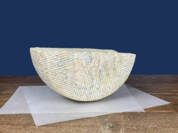 A half wheel of Manchego Curado cheese on greaseproof paper atop wood, with a blue background.