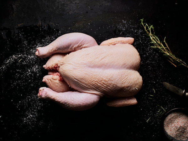 Whole Chicken on a dark surface with salt, herbs, and a knife nearby.